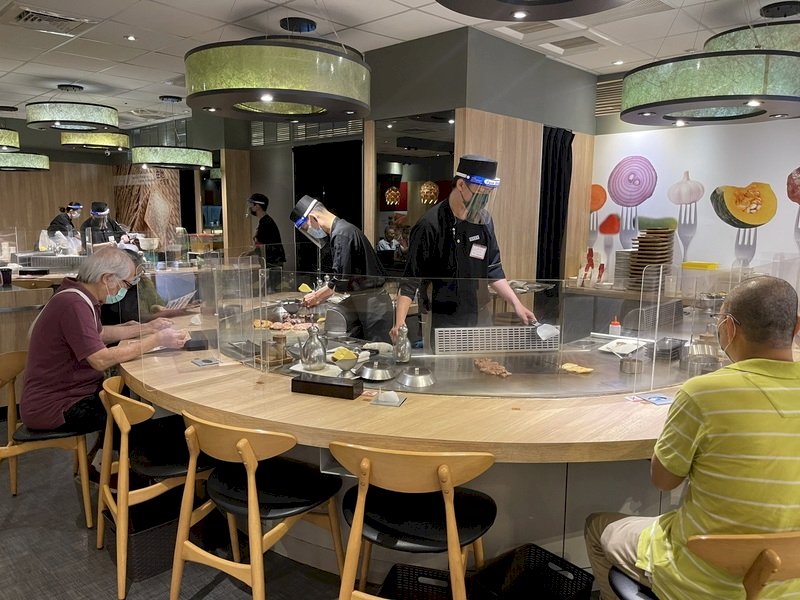 全國疫情警戒降為2級,高雄有條件開放餐廳內用,百貨美食街的鐵板燒業者在桌面架設透明隔板,廚師也都配戴口罩及面罩加強防疫。 (圖:中央社)
